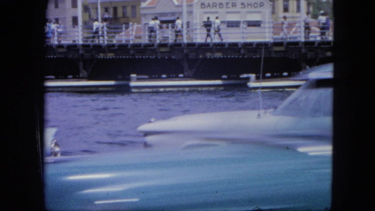 Blue car hood and white car roof by water, with pedestrians and a barber shop sign beyond the railing.