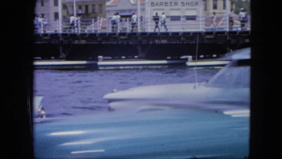 1964 Nassau frame with overlapping car bodies in the foreground, harbor water, pedestrians on the bridge, and a barber shop sign across the waterfront.