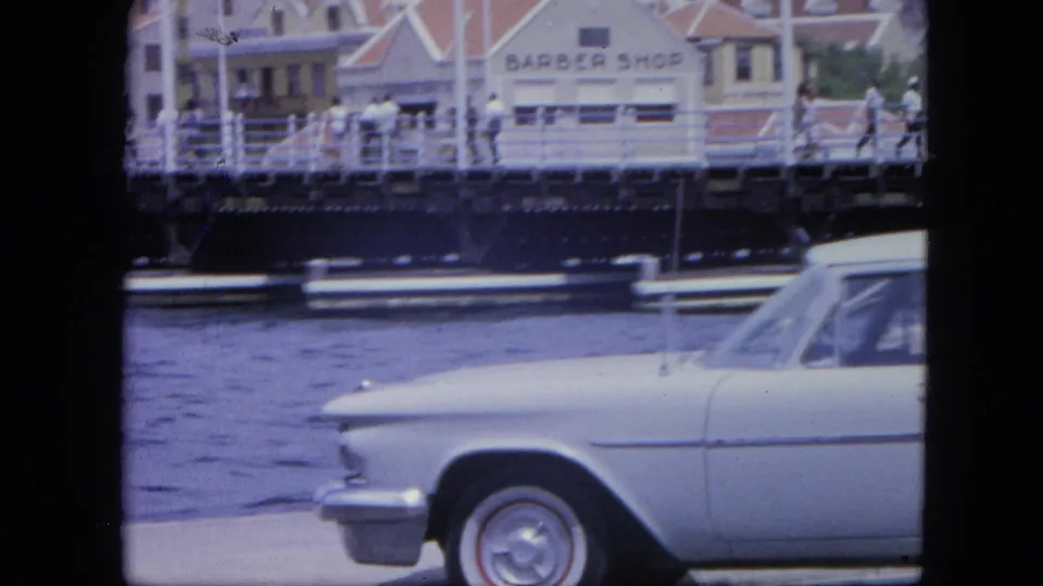 1964 Nassau home-movie frame showing the front half of a white sedan, waterfront water, pedestrians, railing, and a barber shop sign in the background.