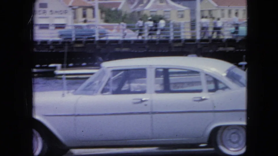 1964 Nassau frame with a white sedan filling the foreground, water and railings behind it, and pedestrians near waterfront buildings.