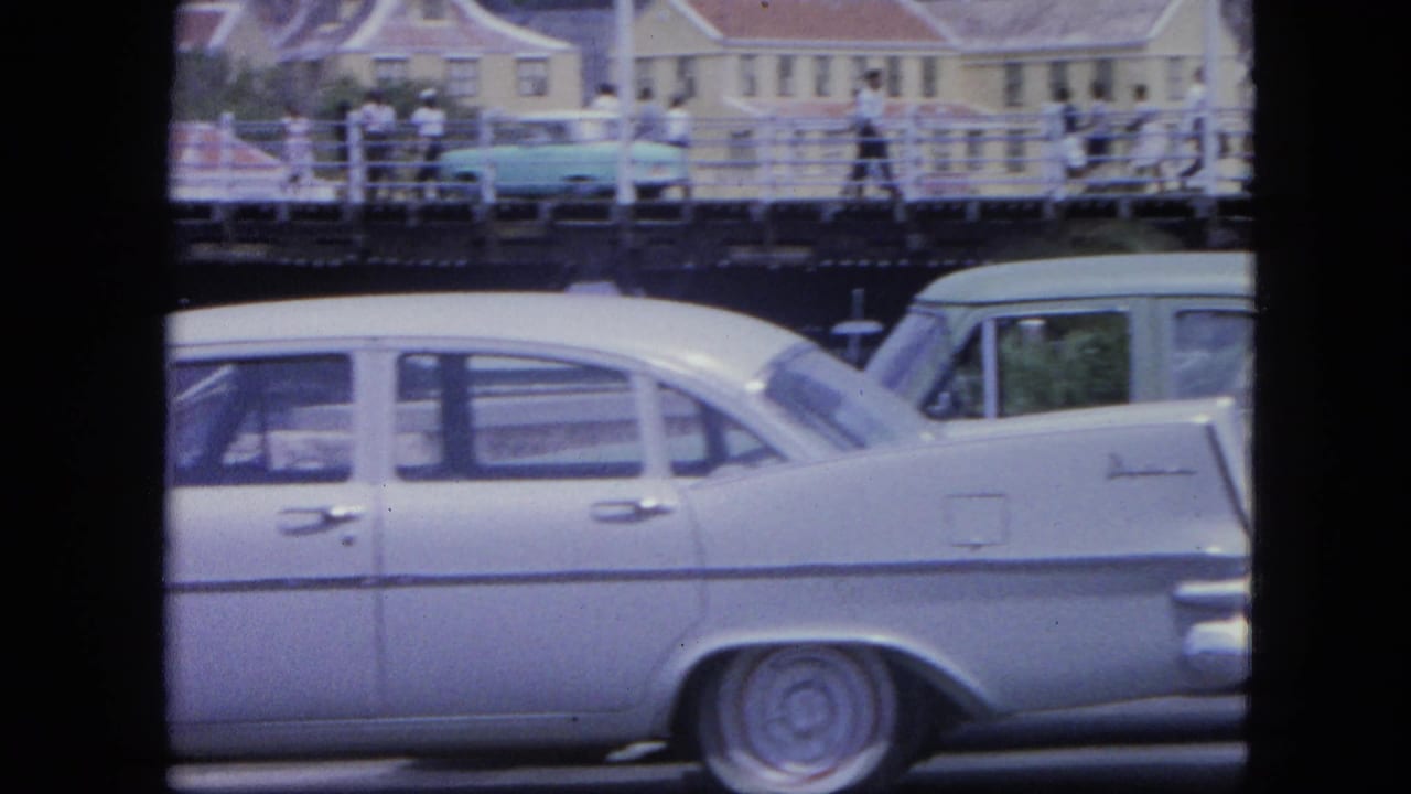 Classic cars pass below a raised walkway with pedestrians in the background.