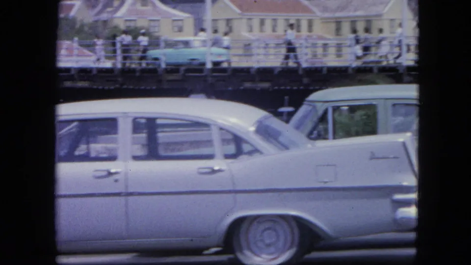 1964 Nassau home-movie frame with classic cars in the foreground and pedestrians crossing a raised walkway behind them.