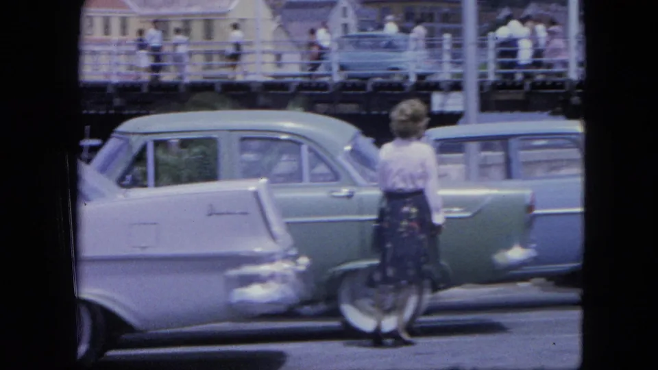 1964 Nassau street frame showing a woman in a patterned skirt beside a green and white station wagon, with pedestrians on a walkway behind.