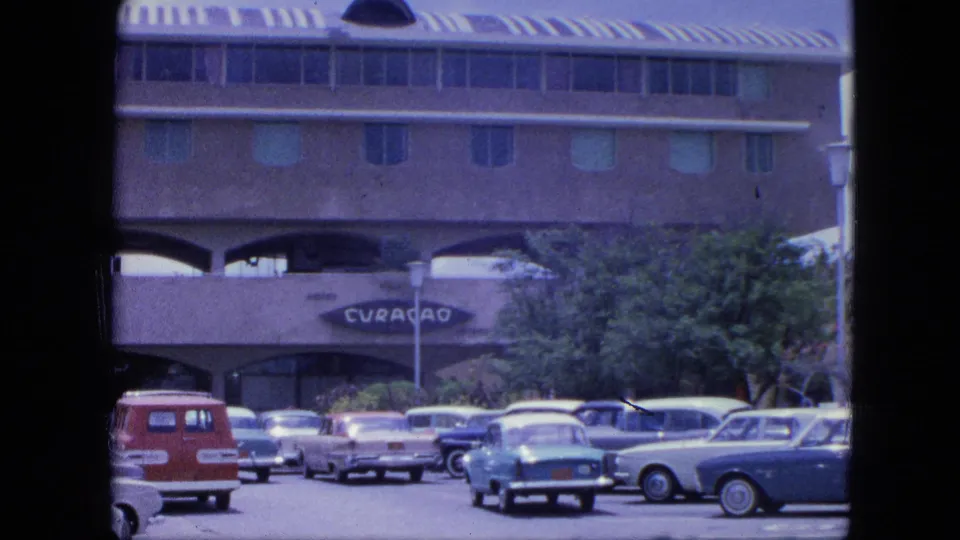 1964 Nassau home-movie view of parked classic cars and a red van in front of a building with a visible CURACAO sign.