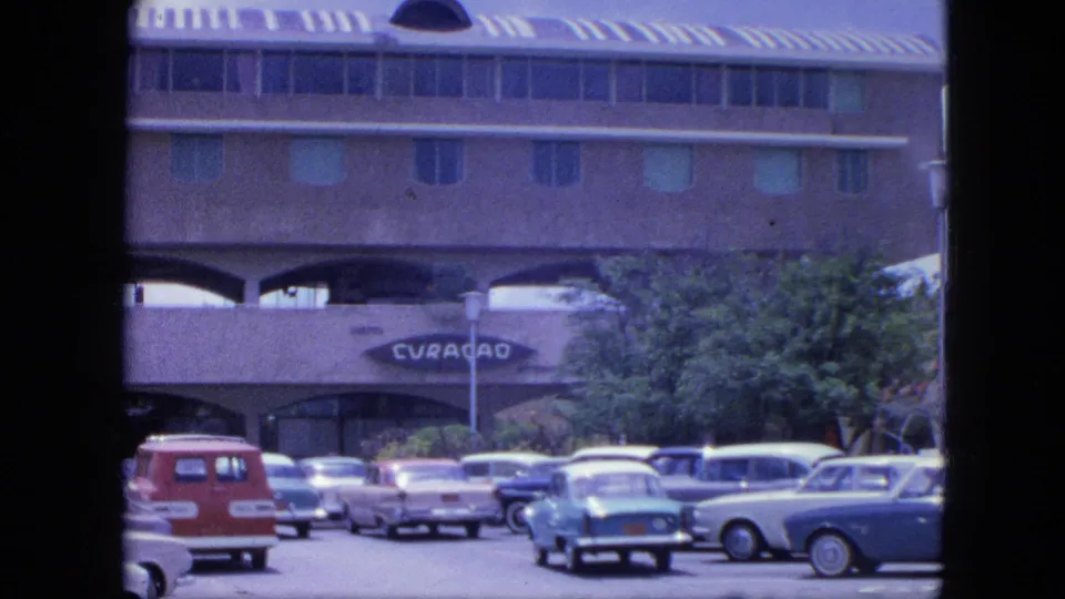 1964 Nassau frame showing a parking area, several classic cars, tropical greenery, and a building entrance marked CURACAO.