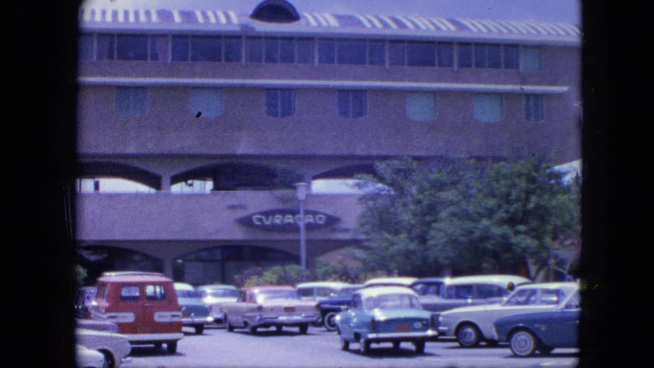 Parking lot with classic cars outside a large arched building with a Curacao sign.