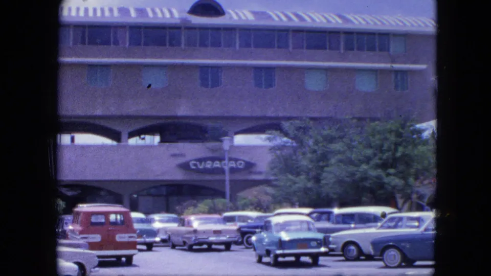 1964 Nassau parking lot scene with classic cars, a red station wagon, trees, and a large arched building marked Curacao.