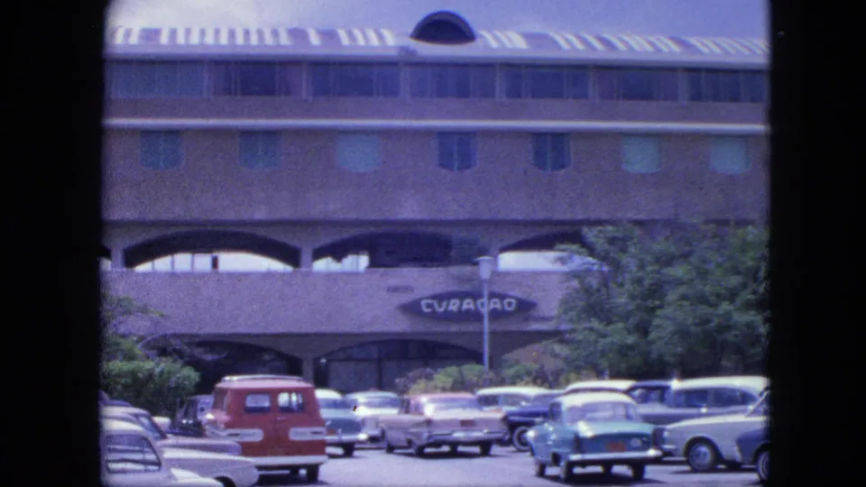 1964 Nassau exterior view of a busy parking lot, midcentury cars, trees, and the Curacao sign on a large arched building.