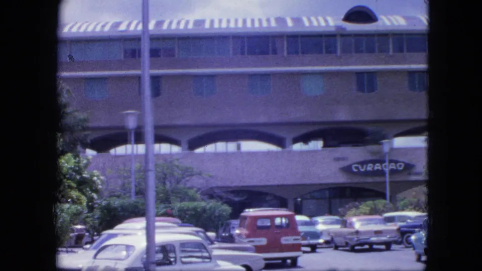 1964 Nassau parking lot view with foreground pole, parked cars, red station wagon, trees, and arched building frontage.