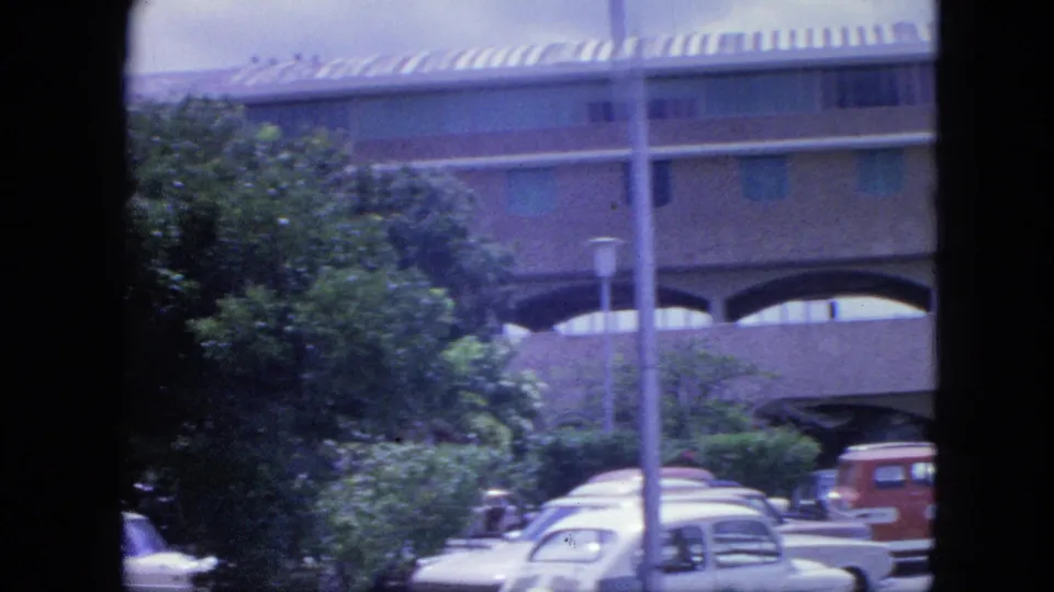1964 Nassau home-movie frame showing parked cars, foliage, a light pole, and the large arched building in soft focus.