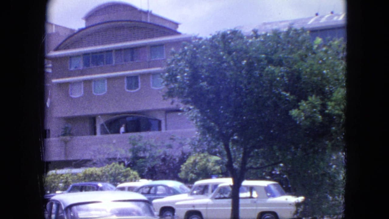 Curved-front building behind trees and parked cars on a sunny street.