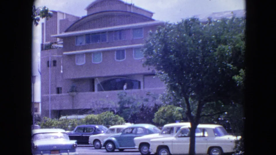 1964 Nassau street frame showing a mid-century building, white and light-colored cars, trees, and roadside landscaping.