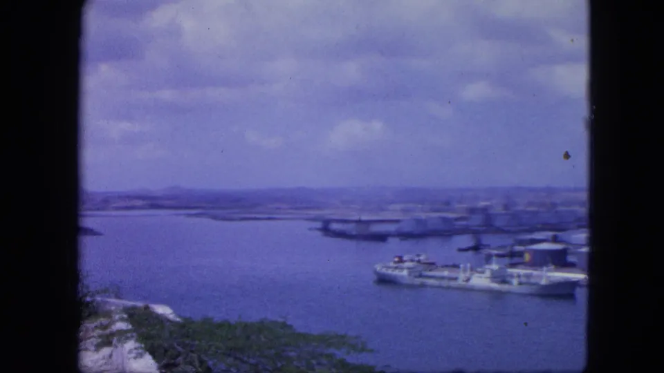 Nassau harbor view in 1964 8mm footage, with blue water, docked boats, shoreline structures, and distant low hills or islands.