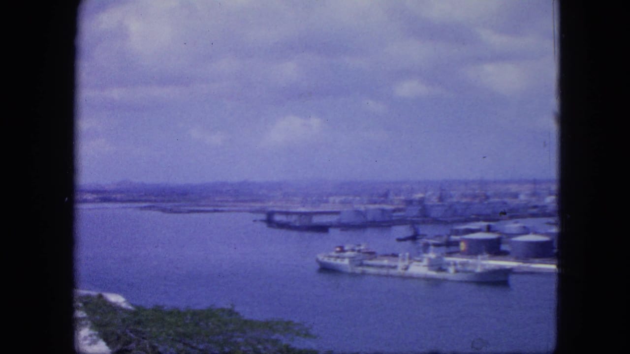 1964 Nassau harbor seen from above, with ships near waterfront tanks and a low skyline under clouds in soft color 8mm footage.