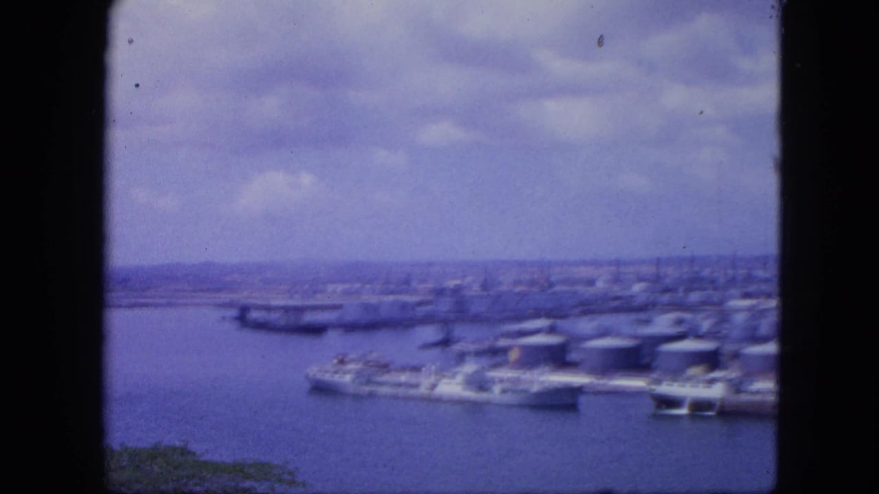 Nassau, Bahamas harbor panorama from 1964, showing vessels beside industrial tanks and a distant city skyline through aged 8mm color film.