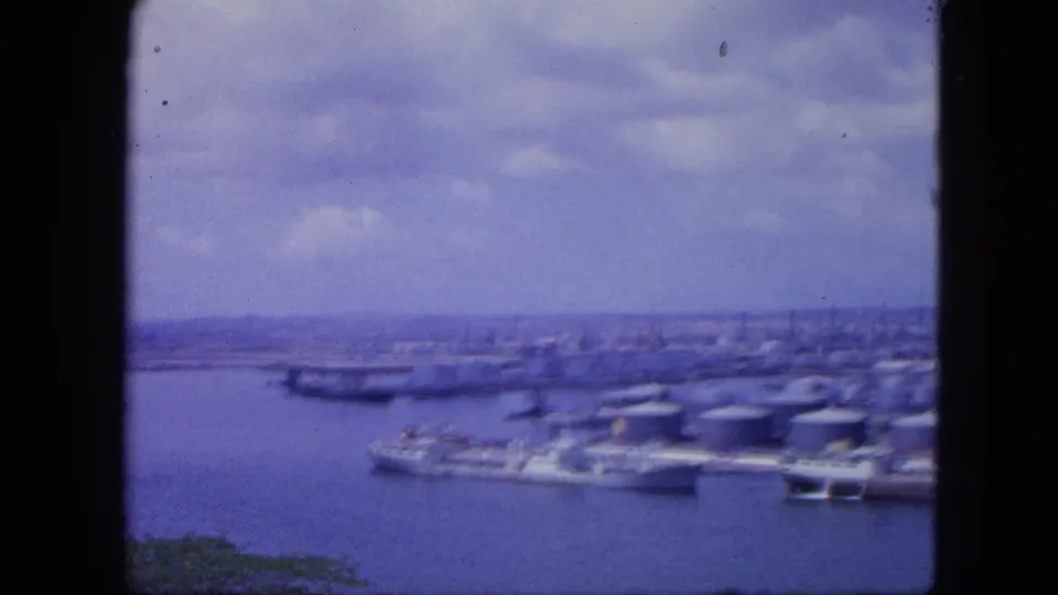 Nassau, Bahamas harbor panorama from 1964, showing vessels beside industrial tanks and a distant city skyline through aged 8mm color film.