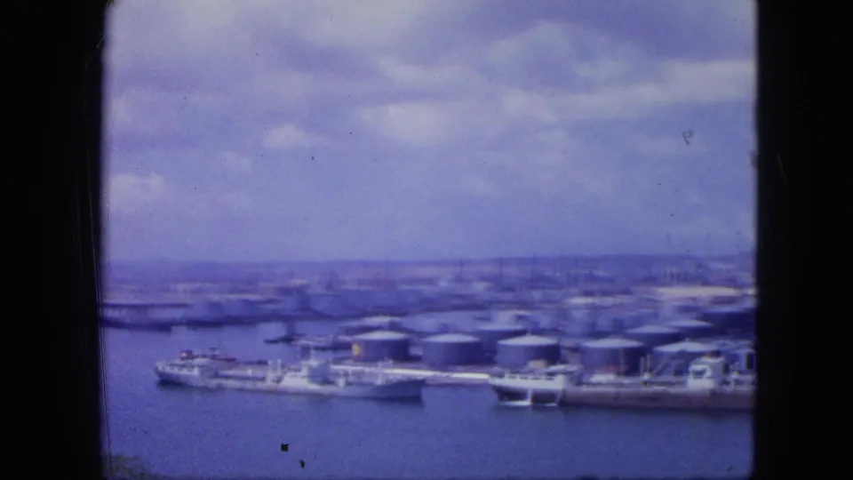 1964 Nassau waterfront view with a long vessel at dock, clustered storage tanks, piers, and distant city buildings in soft 8mm color.