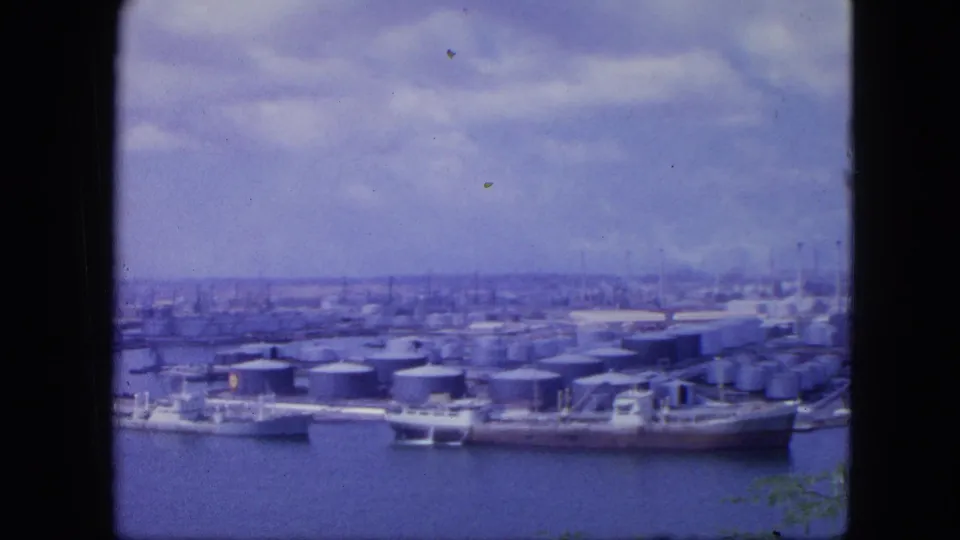 Nassau harbor in 1964 with multiple storage tanks, docked vessels, distant buildings, and a small edge of greenery in vintage 8mm color.