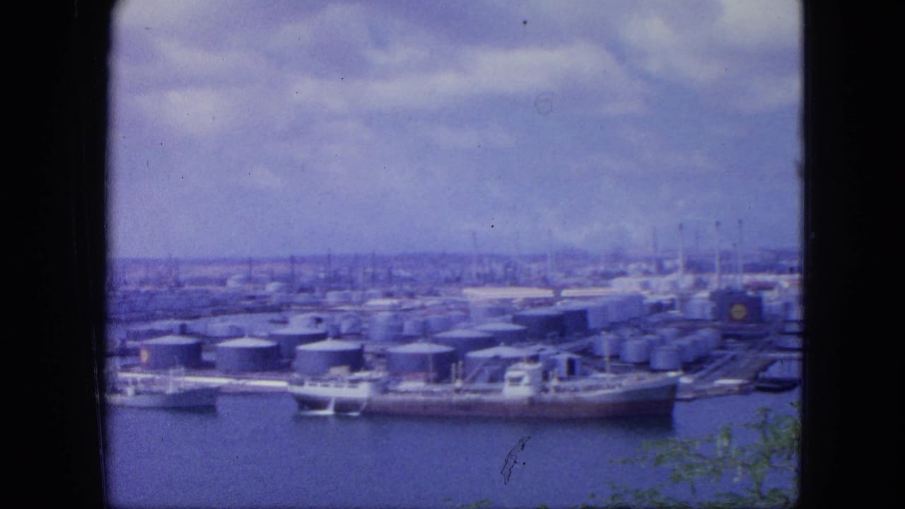 1964 Nassau harbor view with docked tankers, clustered storage tanks, industrial buildings, water, and a cloudy sky.