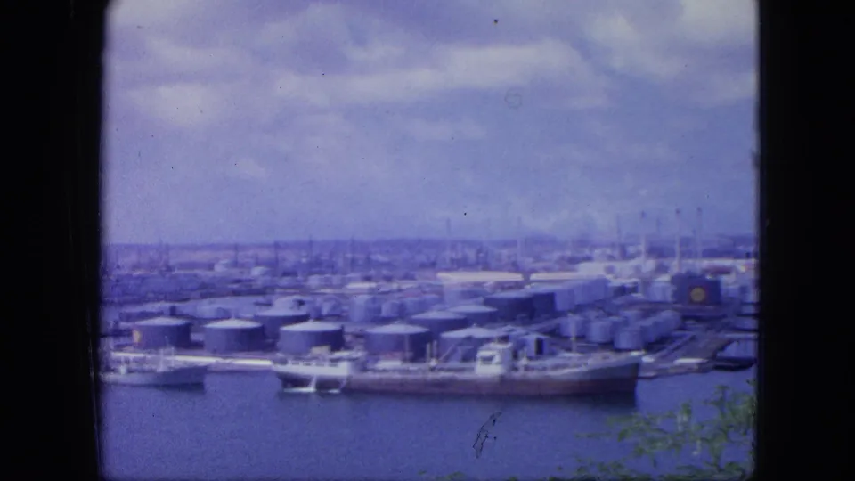 1964 Nassau harbor view with docked tankers, clustered storage tanks, industrial buildings, water, and a cloudy sky.