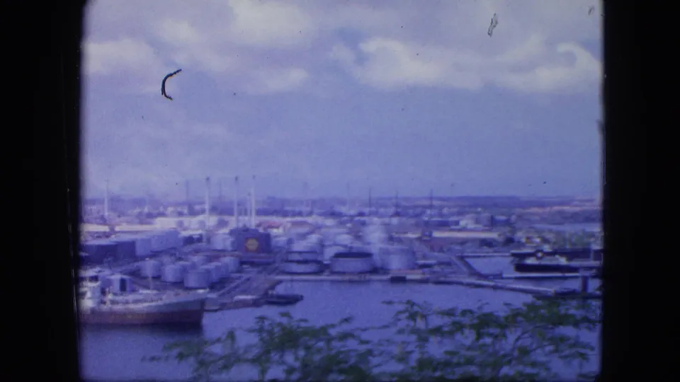 1964 Nassau port panorama with fuel tanks, piers, docked ship edges, cloudy sky, and foliage in the foreground.