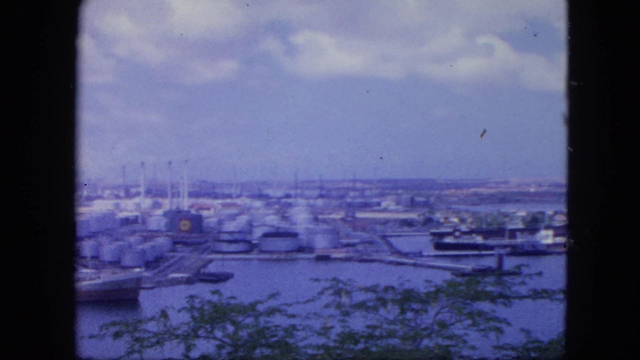 1964 Nassau harbor seen from an elevated viewpoint, with storage tanks, docked vessels, water, clouds, and dark edges framing the 8mm shot.