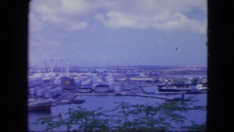 1964 Nassau harbor seen from an elevated viewpoint, with storage tanks, docked vessels, water, clouds, and dark edges framing the 8mm shot.