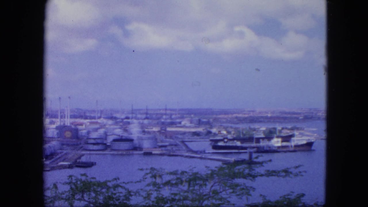 Nassau, Bahamas port waterfront in 1964, showing tank farms, a docked ship, harbor water, distant shoreline, clouds, and dark foreground framing.