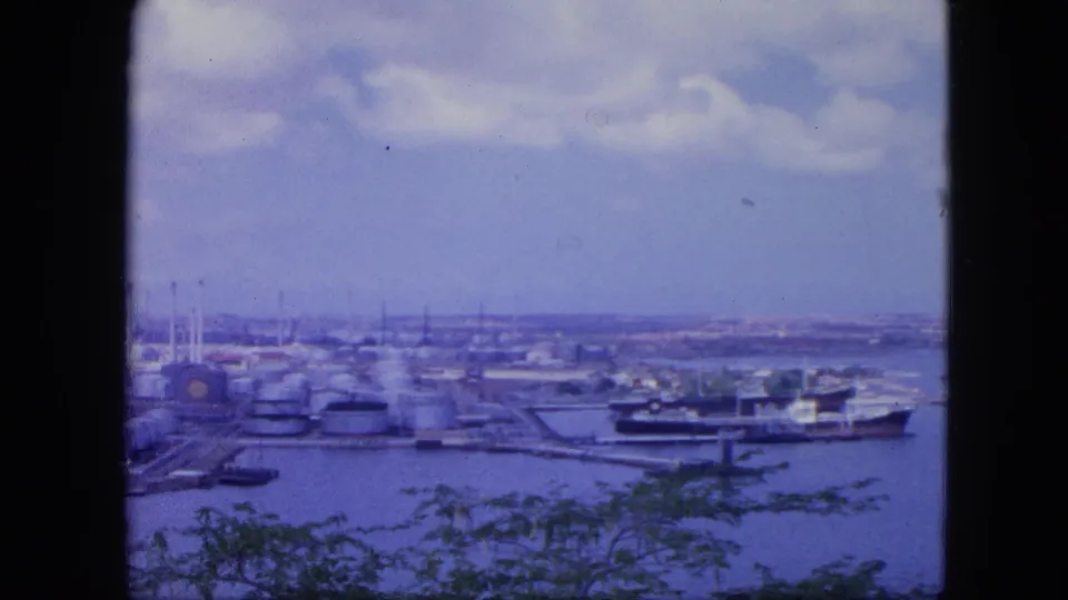 Nassau, Bahamas port waterfront in 1964, showing tank farms, a docked ship, harbor water, distant shoreline, clouds, and dark foreground framing.