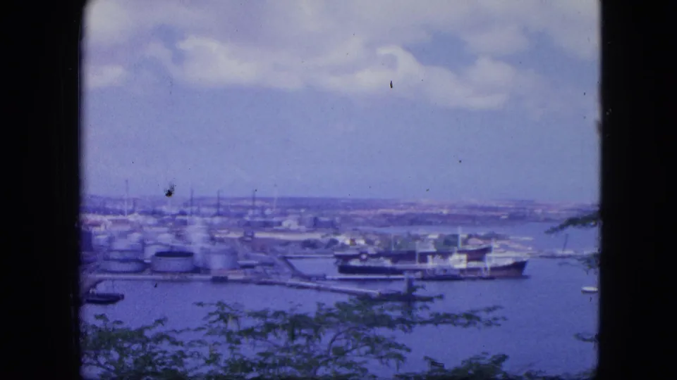 1964 8mm view over Nassau harbor, with a large vessel near storage tanks, low waterfront buildings, distant land, and foreground foliage.