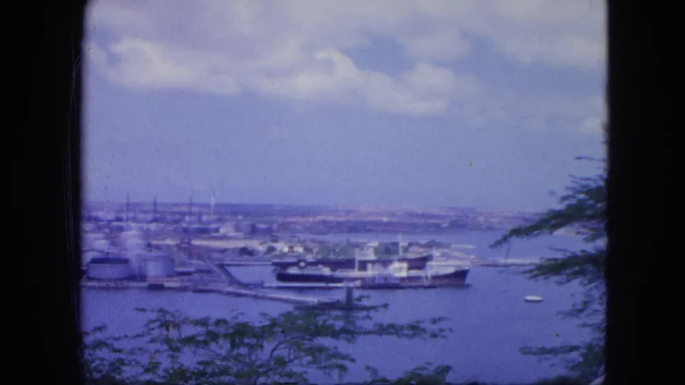 End of the 1964 Nassau harbor pan, showing a docked ship, tank storage, open water, distant shoreline, clouds, and foliage at the frame edge.