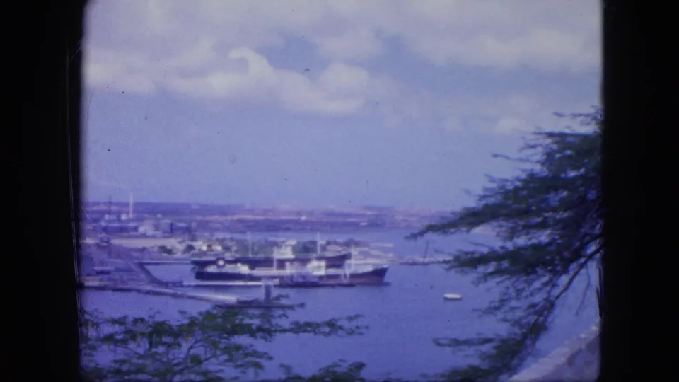Elevated 1964 Nassau harbor frame with a moored ship, small boats, distant port area, clouds, and trees along the edge.