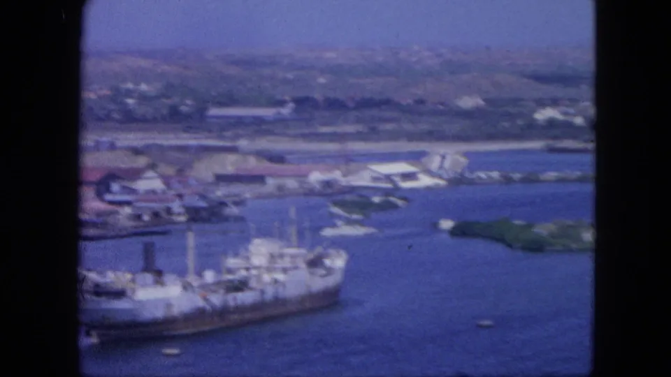 Wide 1964 Nassau harbor frame showing a large docked ship, small boats, waterfront structures, blue water, and distant shoreline.