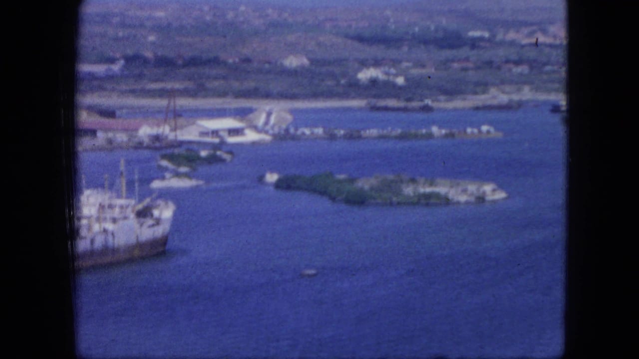 1964 Nassau aerial view showing harbor water, low islands, dock structures, shoreline buildings, and a large vessel at left.