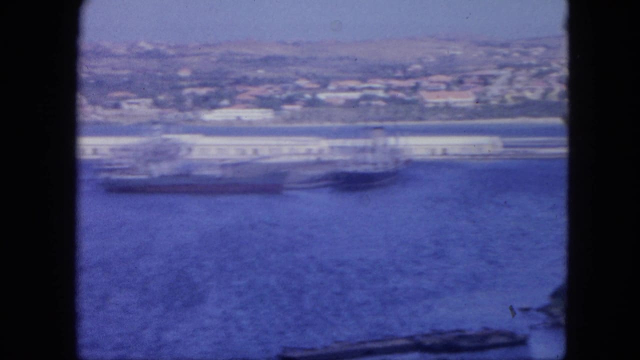 1964 Nassau waterfront with a large vessel at the dock, blue bay water, and hillside buildings in the distance.