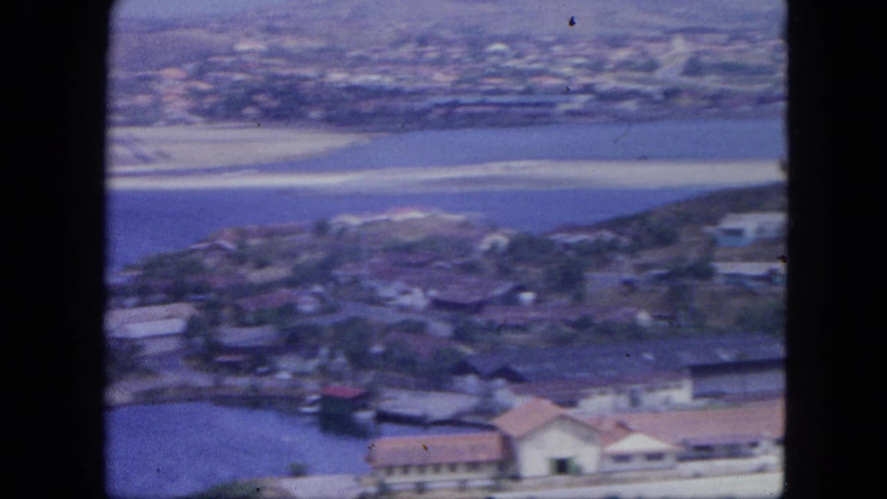 Soft aerial view of shoreline buildings, harbor water, and low hills beyond a coastal town.