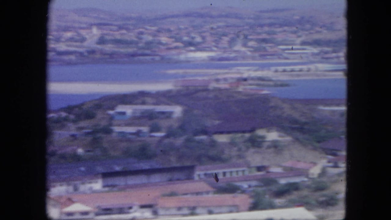 1964 Nassau aerial frame with harbor water, shoreline development, low buildings, and distant coastal town visible through soft 8mm color.