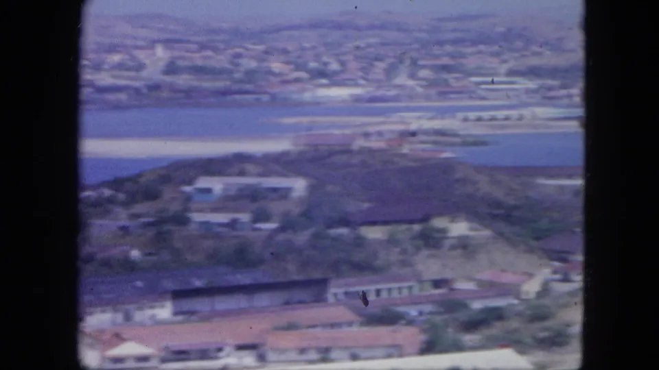1964 Nassau aerial frame with harbor water, shoreline development, low buildings, and distant coastal town visible through soft 8mm color.