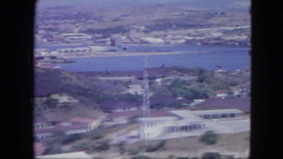 1964 8mm view over Nassau with a communication tower, low buildings, harbor water, shoreline roads, and distant hills.