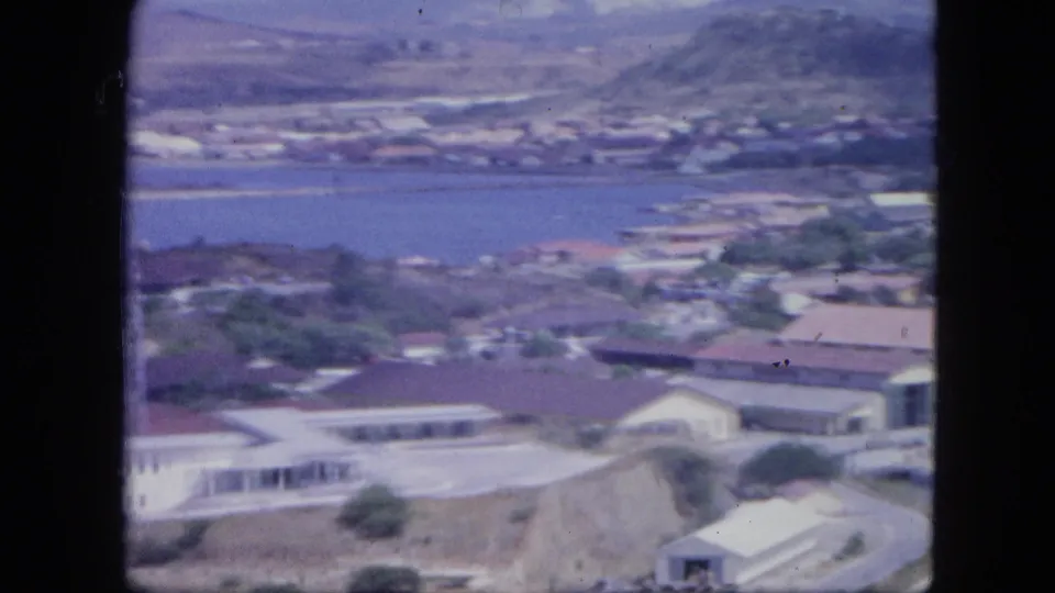1964 Nassau aerial view showing harbor water, hillside buildings, foreground rooftops, roads, and soft small-gauge film texture.