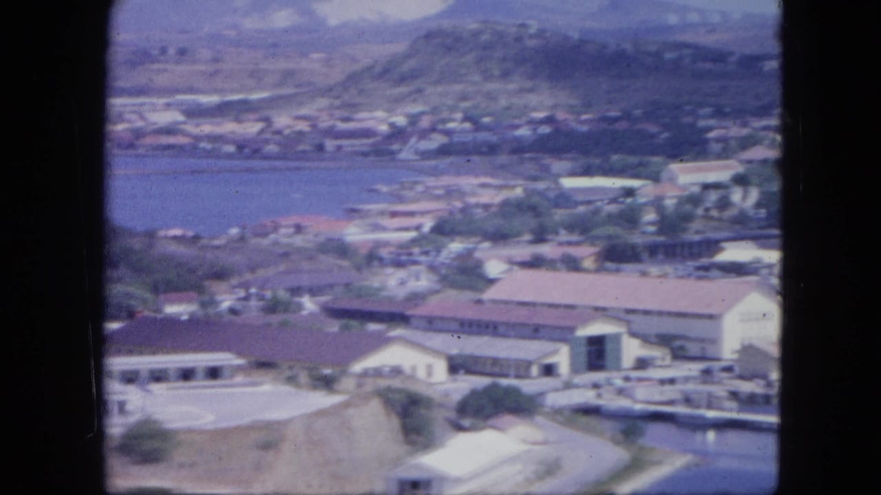 1964 Nassau aerial view showing waterfront structures, red-roofed buildings, harbor water, and low hills beyond.