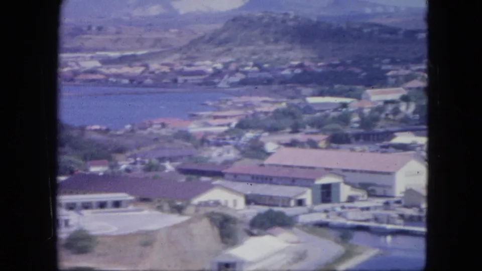 1964 Nassau aerial view showing waterfront structures, red-roofed buildings, harbor water, and low hills beyond.
