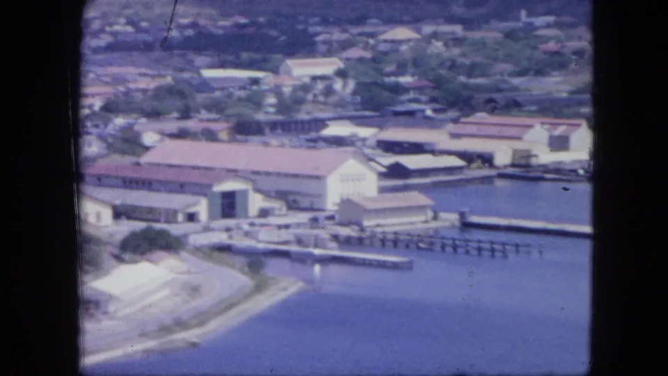 Aerial Nassau harbor view with large waterfront buildings, piers, moored boats or small craft, and hillside town beyond.