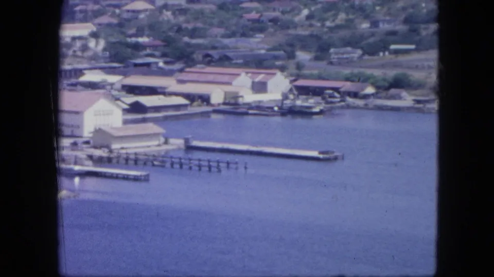 1964 Nassau view looking across blue harbor water toward a pier, shoreline buildings, and low residential hills.