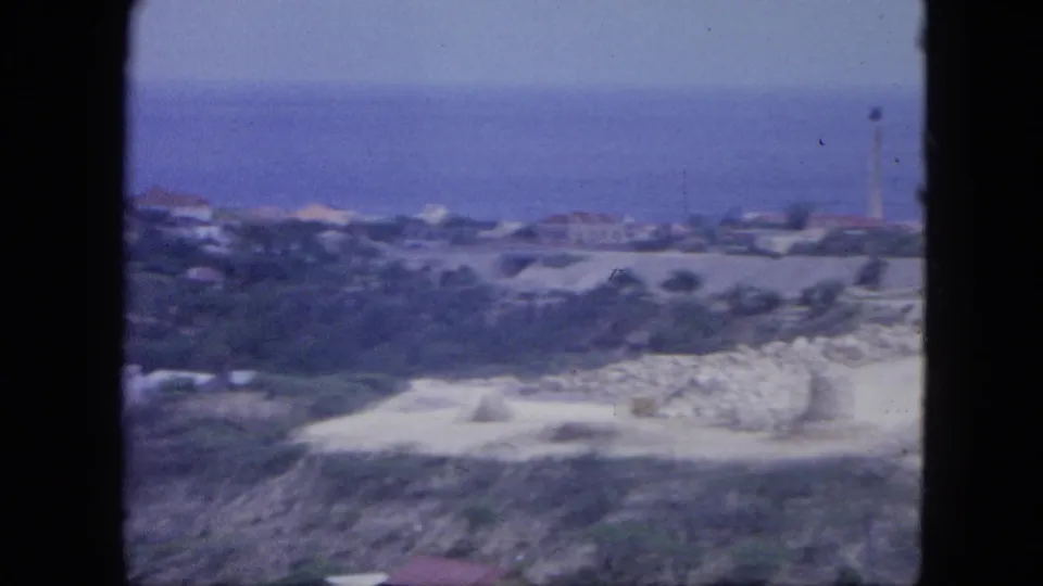 Elevated Nassau coastal view with open ocean beyond low buildings, pale ground, and a distant lighthouse-like tower.