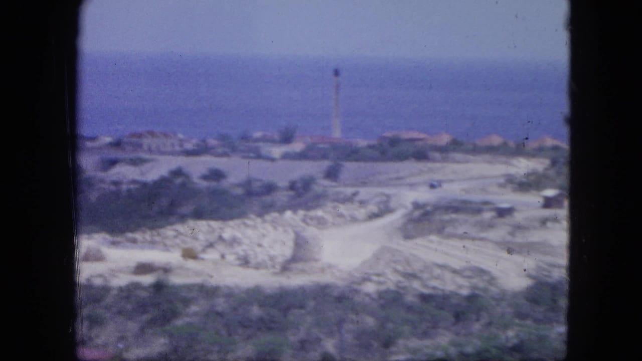 Soft coastal view with winding dirt roads, low buildings, sea horizon, and a distant tower.