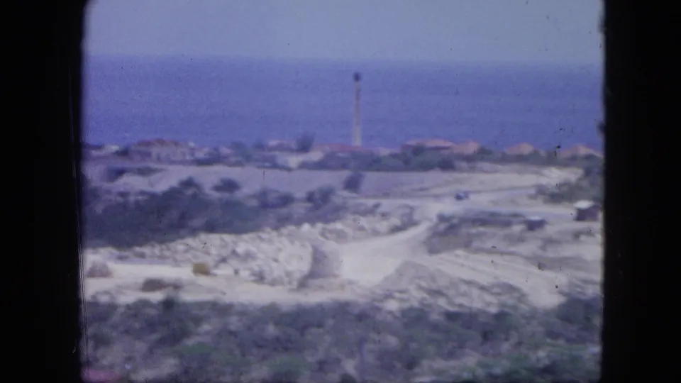 1964 Nassau home-movie view of a coastal area with pale roads, scrubland, low buildings, blue sea, and a distant lighthouse-like tower.