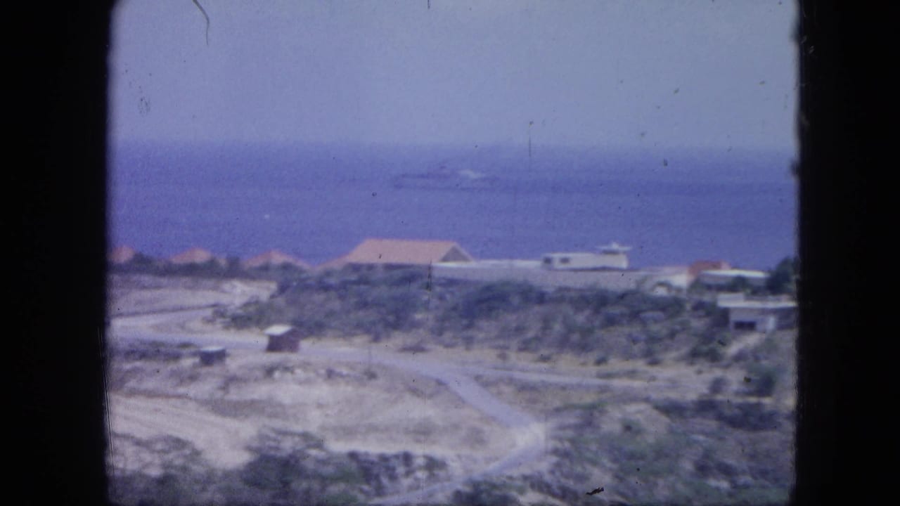 Coastal settlement with blue sea, low houses, rocky ground, and a pale road in the foreground.