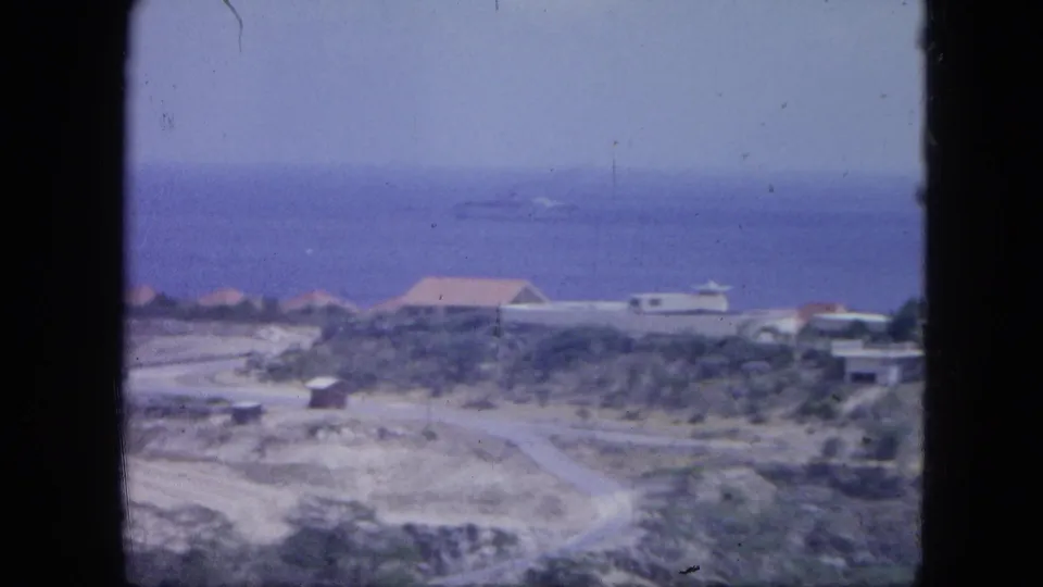 1964 Nassau coastal scene showing low buildings, rocky terrain, a pale road, and the blue sea beyond.