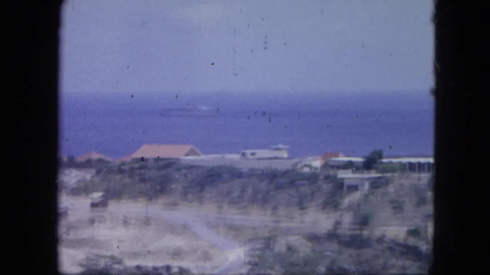 1964 Nassau home-movie frame with low coastal buildings, a road, scrubland, and the sea in soft blue-purple color.